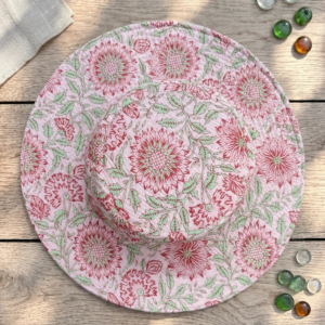 Top-down-view-of-a-light-blush-pink-quilted-wide-brim-sun-hat-featuring-an-intricate-red-and-green-floral-block-print-pattern-resting-on-a-light-colored-wooden-table