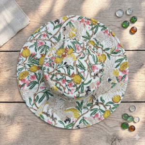 Top-down-view-of-an-off-white-quilted-wide-brim-sun-hat-decorated-with-a-complex-botanical-chintz-pattern-featuring-crested-cockatoos-lemons-and-pink-flowers-placed-on-a-light-wooden-table