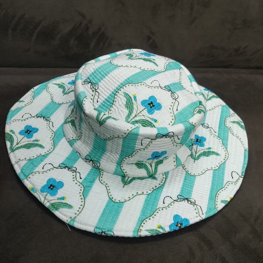 A-white-and-mint-green-vertically-striped-quilted-wide-brim-bucket-hat featuring-vintage-inspired-framed-blue-flowers-with-green-leaves-shown-on-a-dark-fabric-background.